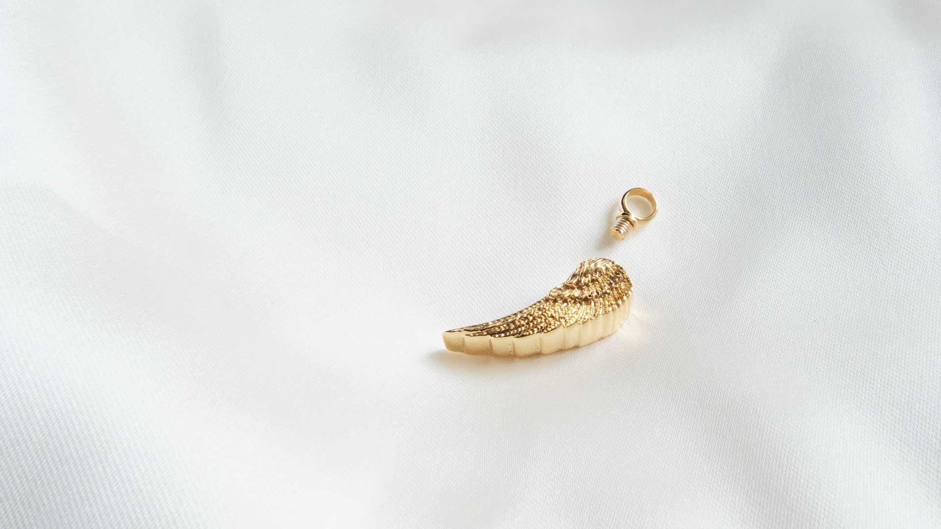 Gold ashes pendant shaped like an angel wing on a light grey background