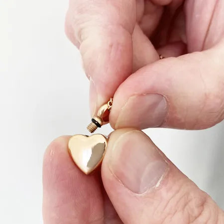 Gold heart-shaped ashes necklace being held between fingers with screw top.