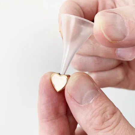 Gold heart-shaped earring held between fingers with a clear plastic funnel tool on a white background